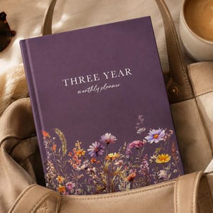 May include: A deep purple hardback planner with the text "THREE YEAR monthly planner" printed in white on the front cover, featuring a colourful botanical illustration of various wildflowers along the bottom edge of the book cover.