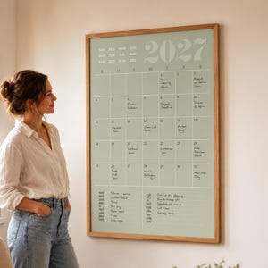 May include: A large wall-mounted 2027 calendar with a sage green background and a light wood frame. The grid displays days of the month with handwritten notes, a menu section, and a notes section, all printed in white text on a sage green surface.