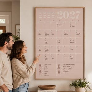 May include: A large wall-mounted 2027 calendar in a light pink hue with a wooden frame. The calendar features a monthly grid with handwritten-style text for dates and events, a dedicated menu section, and a notes column with bulleted list items.