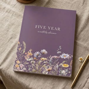 May include: A five-year monthly planner with a purple cover featuring floral artwork. The cover has the text "FIVE YEAR" above "monthly planner" in a script font. A gold pen and a small flower are next to the planner on a neutral-colored surface.