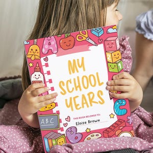 Peut inclure: Un livre rectangulaire intitulé My School Years avec une bordure rose ornée d'illustrations colorées de fournitures scolaires comme des ciseaux, de la colle, des blocs et des livres. La couverture présente une zone centrale blanche avec le texte This book belongs to: Eloise Brown.