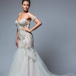 Puede incluir: Una mujer con un vestido de novia blanco con un corpi&ntilde;o ajustado y una falda fluida. El vestido est&aacute; adornado con lentejuelas y abalorios. La mujer est&aacute; de pie sobre un fondo gris.
