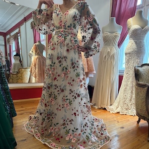 May include: A white maxi dress with a floral pattern. The dress has long sleeves and a v-neckline. The dress is made of a sheer fabric and has a fitted bodice. The dress is displayed on a mannequin against a wooden floor background.