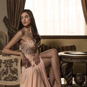 Op de afbeelding: Een vrouw in een roze, met kralen bezette, strapless jurk zit in een vintage stoel in een luxe kamer. De stoel is versierd met houtsnijwerk en bekleed met een bloemenpatroon. De kamer heeft een groot raam met transparante gordijnen, een open haard en een bijzettafel met een donkerhouten blad.