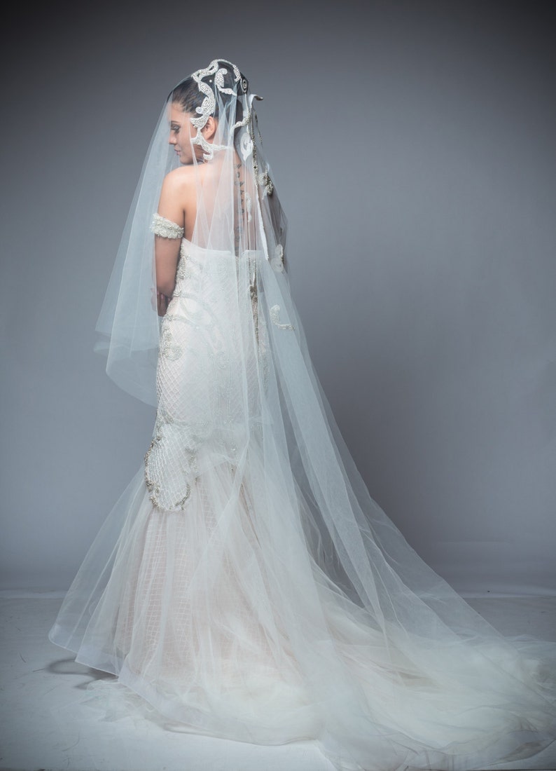 Puede incluir: Una novia con un vestido de novia blanco con un velo largo y un tocado decorativo. El vestido tiene un corpi&ntilde;o ajustado y una falda fluida.