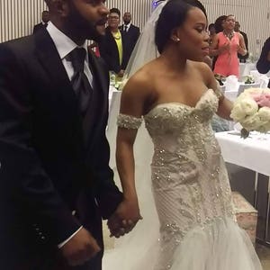 Puede incluir: Una novia y un novio se toman de la mano durante su ceremonia de boda. La novia lleva un vestido blanco sin tirantes con adornos de cuentas y un velo. El novio lleva un traje negro con camisa blanca y corbata negra.