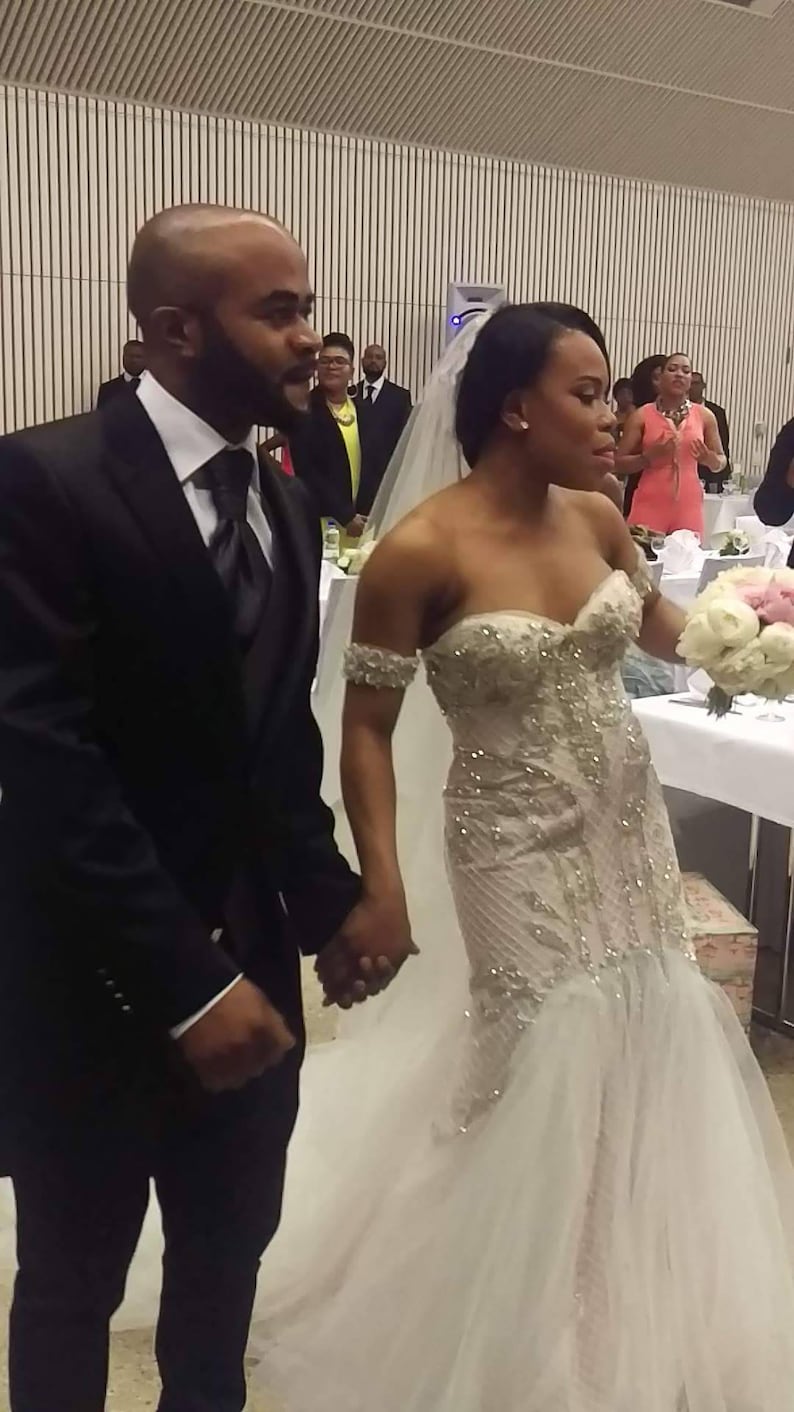 Puede incluir: Una novia y un novio se toman de la mano durante su ceremonia de boda. La novia lleva un vestido blanco sin tirantes con adornos de cuentas y un velo. El novio lleva un traje negro con camisa blanca y corbata negra.