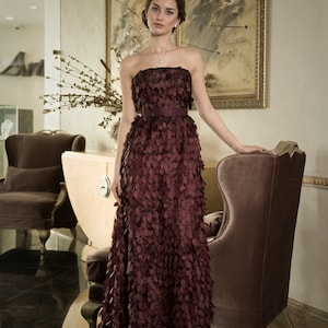 May include: A woman in a burgundy strapless gown with a delicate floral pattern. The gown is made of a soft, flowing fabric and has a fitted bodice. The woman is standing in a room with a plush armchair and a painting on the wall.