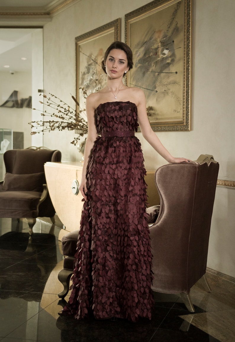 May include: A woman in a burgundy strapless gown with a delicate floral pattern. The gown is made of a soft, flowing fabric and has a fitted bodice. The woman is standing in a room with a plush armchair and a painting on the wall.