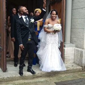 Puede incluir: Una novia y un novio saliendo de una iglesia despu&eacute;s de su ceremonia de boda. La novia lleva un vestido blanco sin tirantes con un velo largo y un ramo de flores blancas. El novio lleva un traje negro con una camisa blanca y una corbata negra. La pareja sonr&iacute;e y est&aacute; feliz.