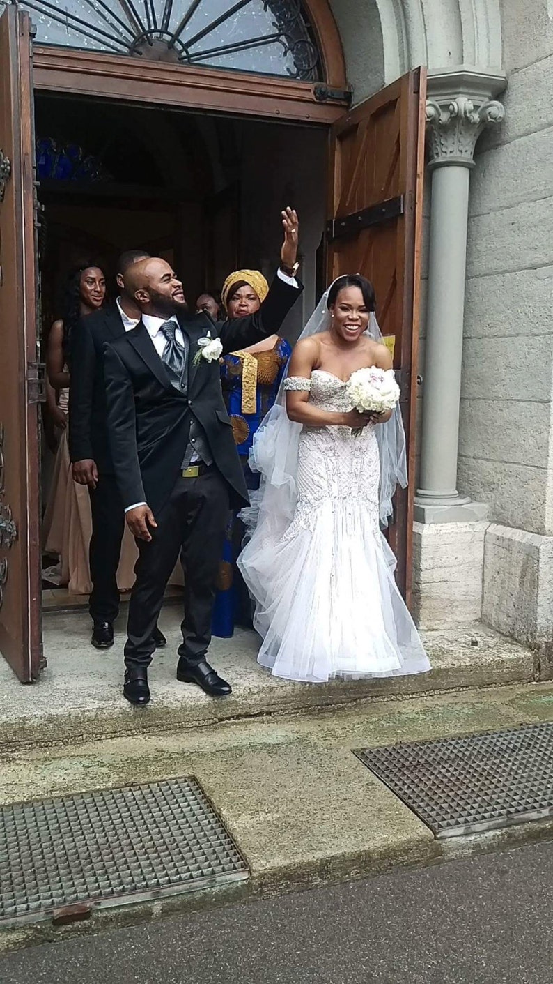 Puede incluir: Una novia y un novio saliendo de una iglesia despu&eacute;s de su ceremonia de boda. La novia lleva un vestido blanco sin tirantes con un velo largo y un ramo de flores blancas. El novio lleva un traje negro con una camisa blanca y una corbata negra. La pareja sonr&iacute;e y est&aacute; feliz.