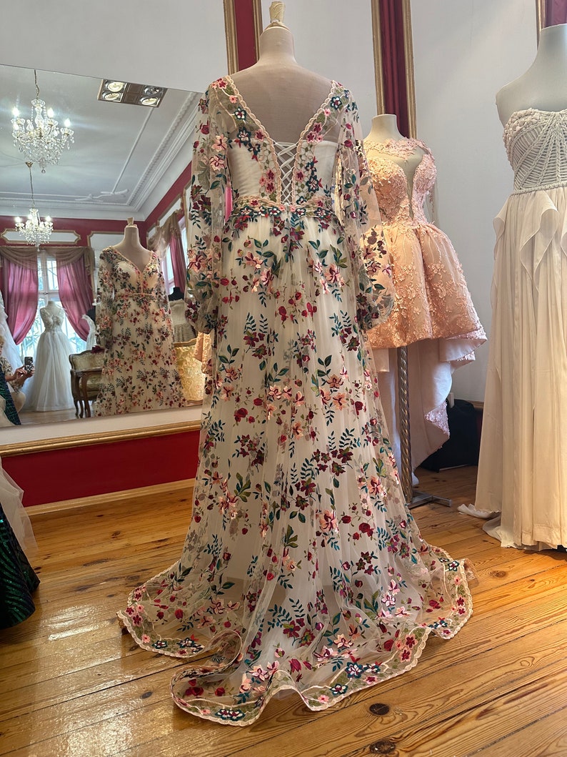 May include: A white long-sleeved gown with a floral pattern. The gown has a v-neckline and a lace-up back. The gown is made of a sheer fabric and has a flowing skirt.