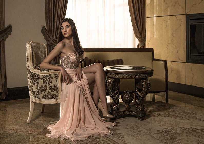 Op de afbeelding: Een vrouw in een roze, met kralen bezette, strapless jurk zit in een vintage stoel in een luxe kamer. De stoel is versierd met houtsnijwerk en bekleed met een bloemenpatroon. De kamer heeft een groot raam met transparante gordijnen, een open haard en een bijzettafel met een donkerhouten blad.