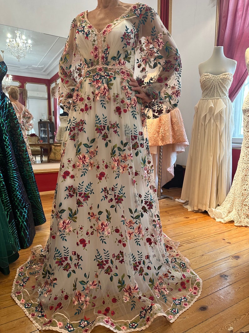 May include: A white maxi dress with a floral pattern. The dress has a v-neckline, long sleeves, and a fitted bodice. The skirt is full and flowing. The dress is made of a sheer fabric with embroidered flowers in shades of pink, red, green, and blue.