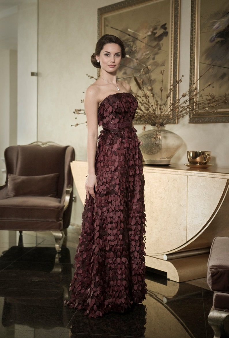 May include: A woman in a burgundy strapless gown with a floral pattern. The gown is made of a soft, flowing fabric and has a fitted bodice. The woman is standing in a room with a gold and white dresser in the background.