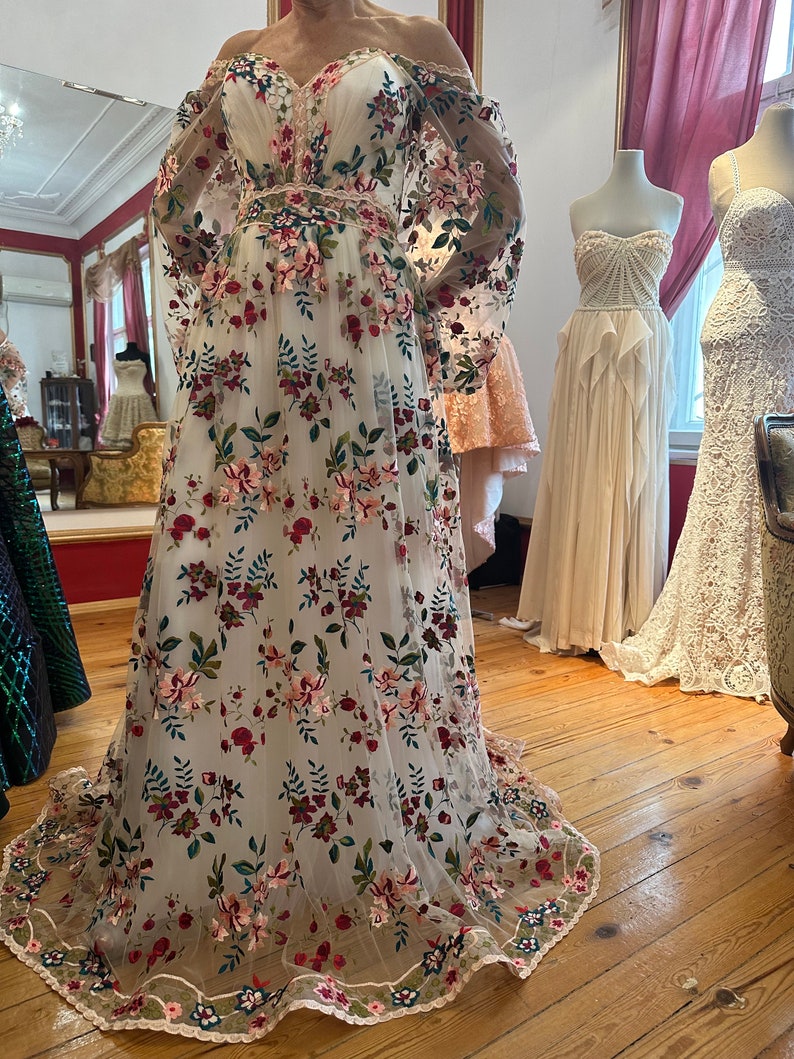 May include: A white off-the-shoulder gown with long sleeves and a floral embroidery pattern. The embroidery features pink, red, and green flowers on a white mesh fabric. The gown has a fitted bodice and a flowing skirt. The gown is displayed on a mannequin in a room with wooden floors and a white wall.
