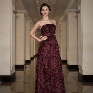May include: A woman in a long, strapless, burgundy gown with a textured, petal-like design. The gown is fitted at the waist and flows out to the floor. She is standing in a hallway with white walls and a patterned ceiling.