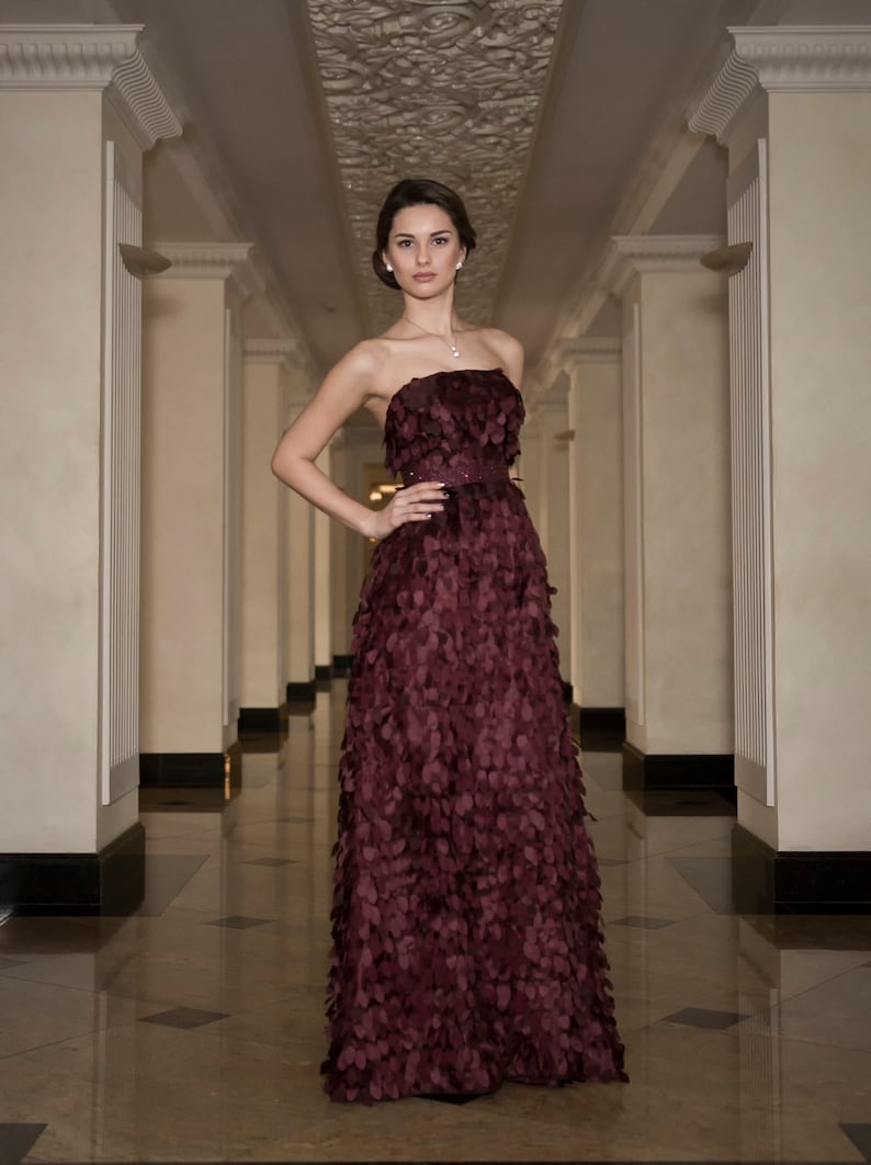 May include: A woman in a long, strapless, burgundy gown with a textured, petal-like design. The gown is fitted at the waist and flows out to the floor. She is standing in a hallway with white walls and a patterned ceiling.