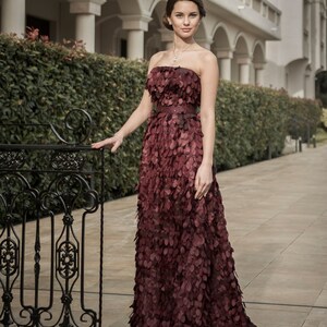 May include: A woman in a burgundy strapless gown with a layered, petal-like design. She is standing in front of a wrought iron gate and a building with white columns.
