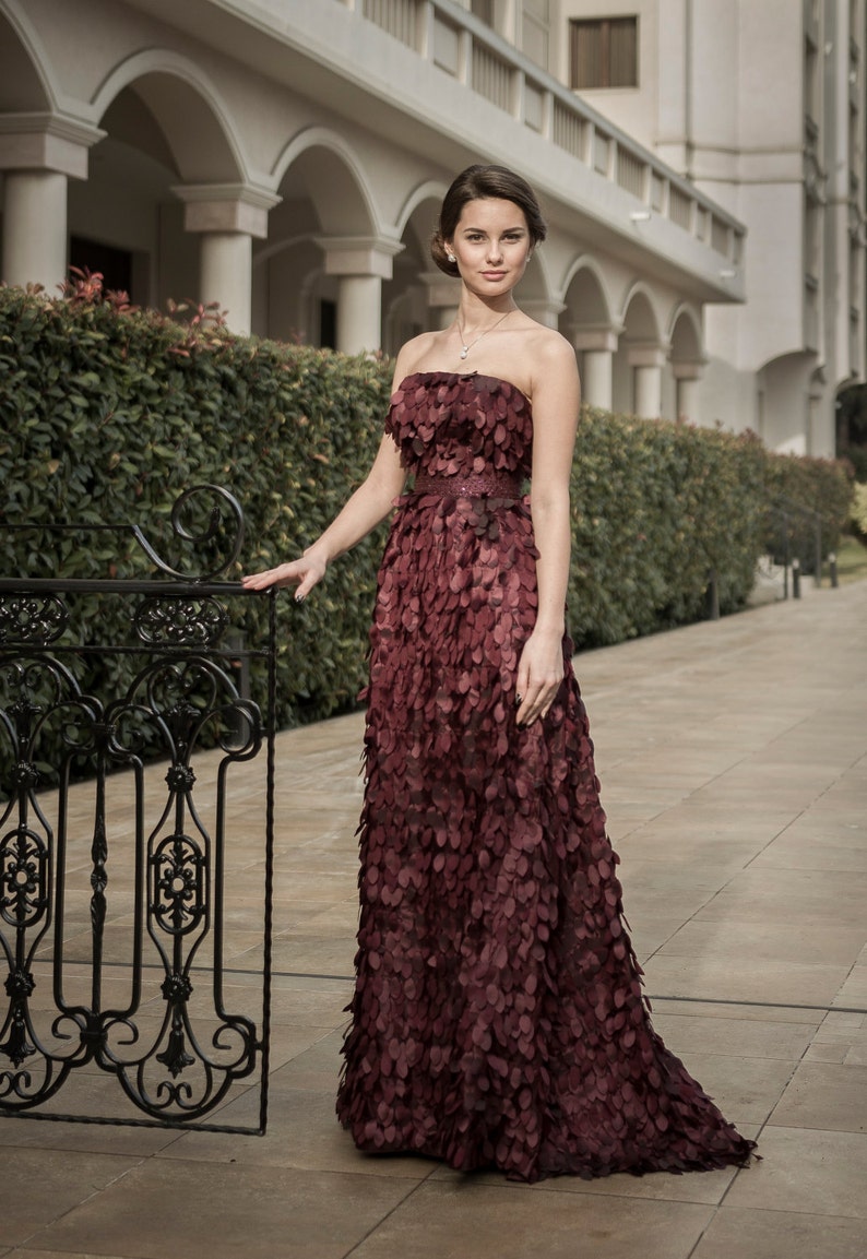 May include: A woman in a burgundy strapless gown with a layered, petal-like design. She is standing in front of a wrought iron gate and a building with white columns.