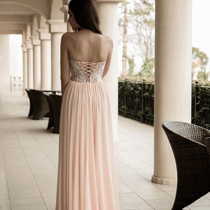 Op de afbeelding: Een vrouw draagt een strapless, lichtroze, lange jurk met een witte kanten bustier en een corset-achtige rug. De jurk is zwierig en heeft een zachte, romantische uitstraling. De vrouw staat in een gang met witte kolommen en uitzicht op bomen op de achtergrond.