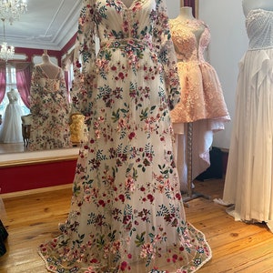 May include: A white maxi dress with a floral pattern. The dress has long sleeves and a v-neckline. The dress is made of a sheer fabric and has a fitted bodice. The dress is embroidered with colorful flowers and leaves. The dress has a lace trim at the hem.