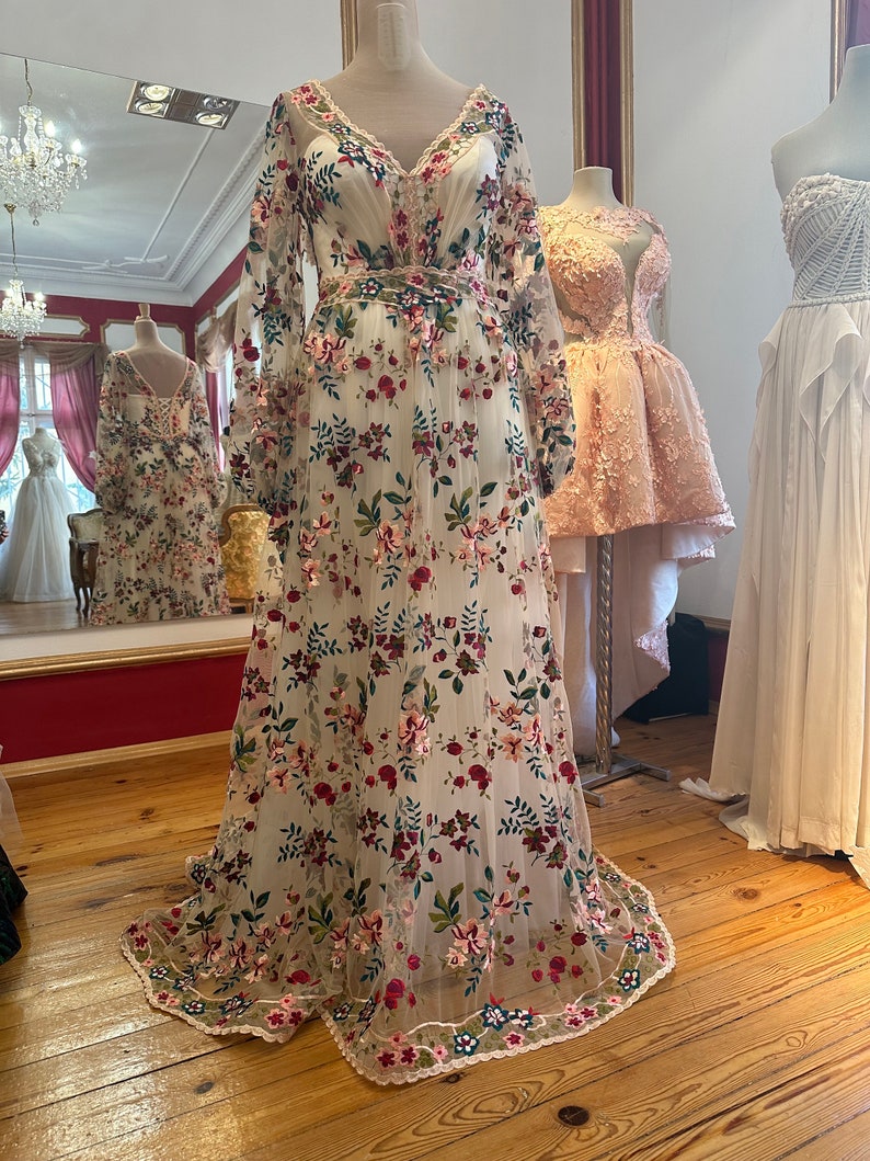 May include: A white maxi dress with a floral pattern. The dress has long sleeves and a v-neckline. The dress is made of a sheer fabric and has a fitted bodice. The dress is embroidered with colorful flowers and leaves. The dress has a lace trim at the hem.