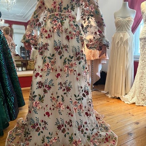 May include: A white off-the-shoulder maxi dress with long sleeves and a floral embroidery pattern in shades of red, pink, green, and brown. The dress has a fitted bodice and a flowing skirt.