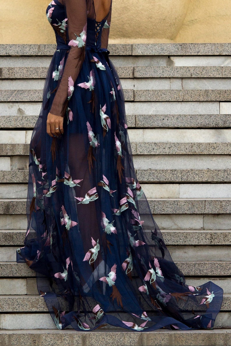 May include: A long, navy blue tulle gown with a sheer bodice and embroidered floral designs. The gown has a fitted bodice and a flowing skirt.