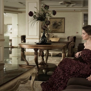May include: A woman in a burgundy dress sits on a couch in a luxurious living room. The room features a large table with a vase of flowers, a large window, and a chandelier.