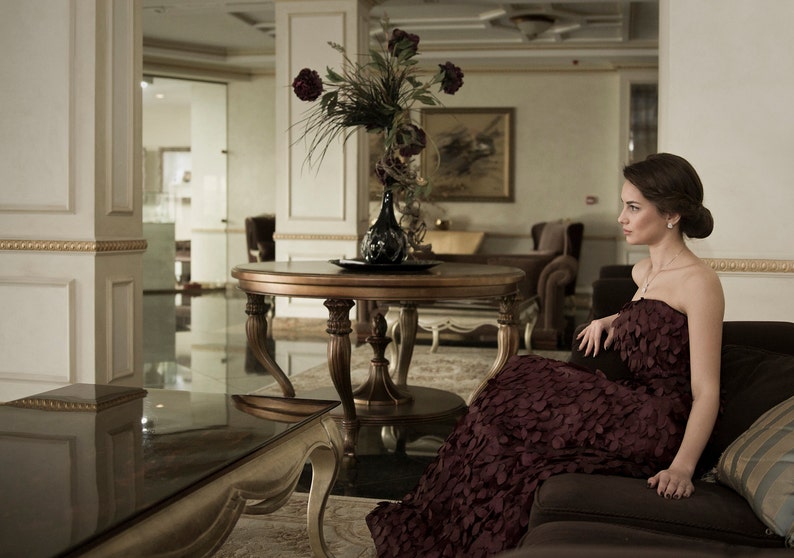 May include: A woman in a burgundy dress sits on a couch in a luxurious living room. The room features a large table with a vase of flowers, a large window, and a chandelier.