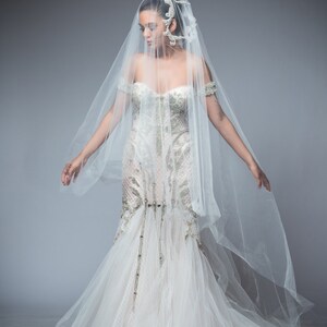 Puede incluir: Una novia con un vestido de novia blanco con un velo largo y una diadema de cuentas. El vestido tiene un corpi&ntilde;o ajustado y una falda fluida.
