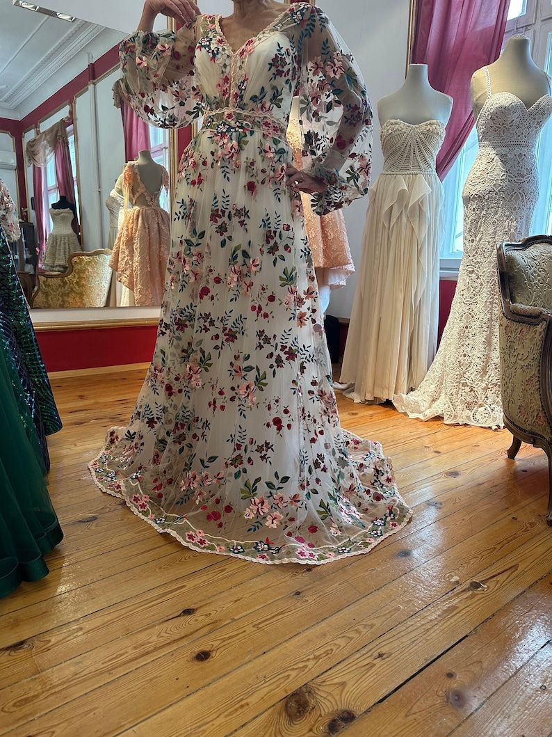 May include: A white maxi dress with a floral pattern. The dress has long sleeves and a v-neckline. The dress is made of a sheer fabric and has a fitted bodice. The dress is displayed on a mannequin against a wooden floor background.