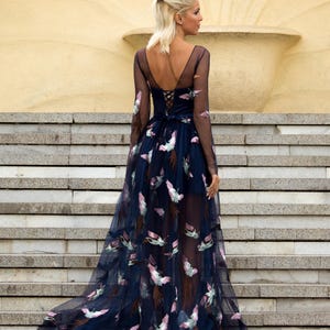 May include: A woman wearing a long, navy blue tulle dress with a floral pattern. The dress has a low back and a lace-up detail. The woman is standing on a set of stairs.