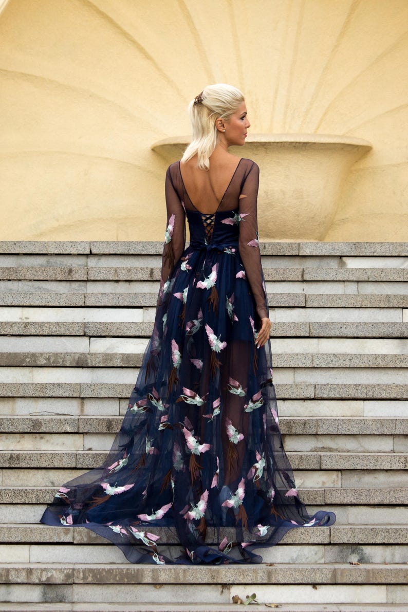 May include: A woman wearing a long, navy blue tulle dress with a floral pattern. The dress has a low back and a lace-up detail. The woman is standing on a set of stairs.