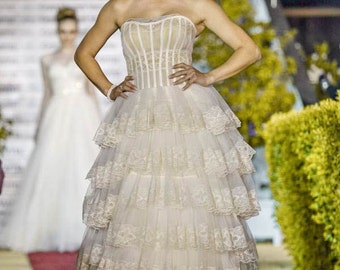 May include: A woman in a white strapless wedding dress with a layered tulle skirt and lace trim. The dress has a fitted bodice with vertical seams. The woman is standing on a red carpet with a green bush in the background.