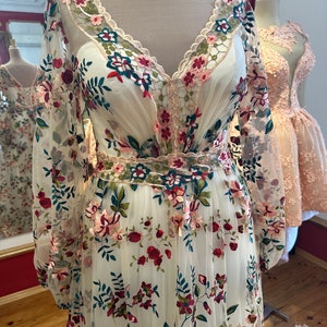 May include: A white maxi dress with a v-neckline and long sleeves. The dress is covered in a floral embroidery pattern with pink, red, green, and blue flowers. The dress has a sheer fabric and a fitted bodice.