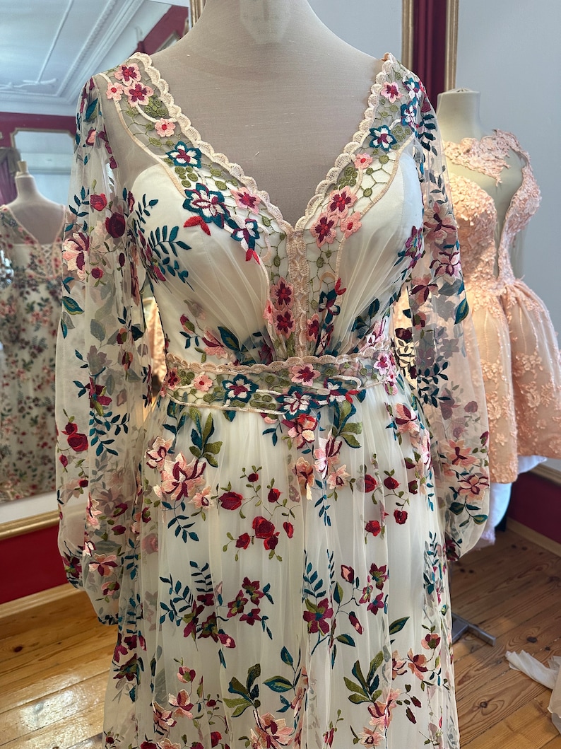 May include: A white maxi dress with a v-neckline and long sleeves. The dress is covered in a floral embroidery pattern with pink, red, green, and blue flowers. The dress has a sheer fabric and a fitted bodice.