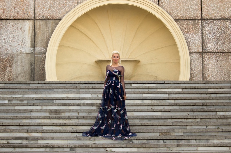 May include: A woman wearing a long, dark blue dress with a floral pattern stands on a set of concrete stairs in front of a large, circular, cream-colored structure.