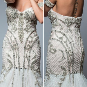 Puede incluir: Un vestido de novia blanco con un corpi&ntilde;o ajustado y una falda fluida. El corpi&ntilde;o est&aacute; cubierto de abalorios y bordados intrincados. La falda est&aacute; hecha de capas de tul y tiene una cola.