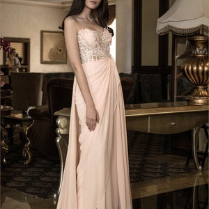 Op de afbeelding: Een vrouw die een strapless, roze jurk draagt met een kralenversierd lijfje en een zwierige rok. De jurk heeft een gedrapeerd ontwerp en is gestyled met een zijsplit. De vrouw staat in een kamer met een gouden lamp en een houten kast op de achtergrond.