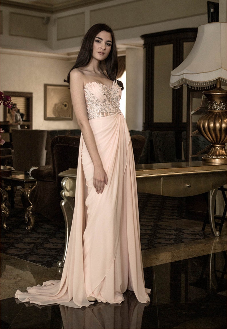 Op de afbeelding: Een vrouw die een strapless, roze jurk draagt met een kralenversierd lijfje en een zwierige rok. De jurk heeft een gedrapeerd ontwerp en is gestyled met een zijsplit. De vrouw staat in een kamer met een gouden lamp en een houten kast op de achtergrond.