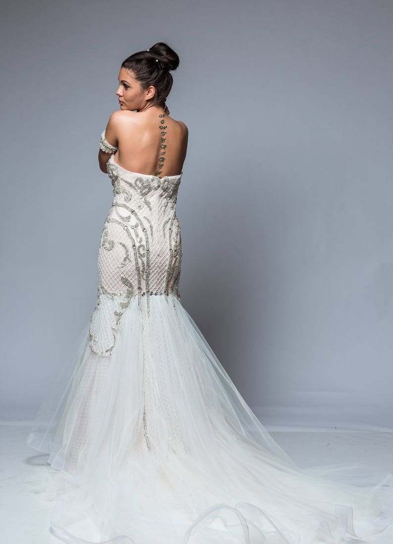 Puede incluir: Una mujer con un vestido de novia blanco con un corpi&ntilde;o de cuentas y una larga cola. El vestido tiene una silueta ajustada y un escote de coraz&oacute;n. La mujer est&aacute; de pie de espaldas a la c&aacute;mara, y su cabello est&aacute; recogido en un mo&ntilde;o.