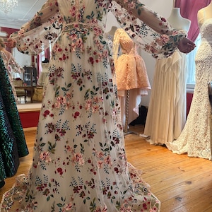 May include: A white maxi dress with long sleeves and a floral pattern. The dress is made of a sheer fabric and has a fitted bodice and a flowing skirt. The flowers are embroidered in shades of pink, red, green, and blue.