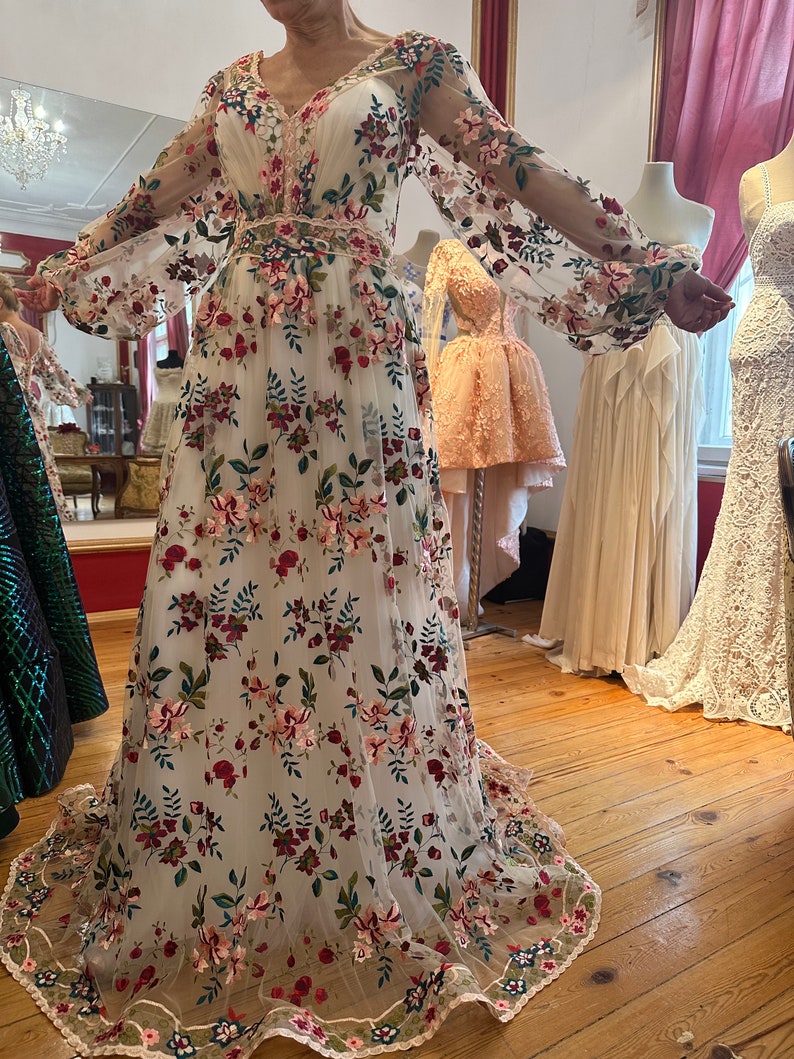 May include: A white maxi dress with long sleeves and a floral pattern. The dress is made of a sheer fabric and has a fitted bodice and a flowing skirt. The flowers are embroidered in shades of pink, red, green, and blue.