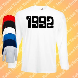 Op de afbeelding: Wit shirt met lange mouwen met het jaar "1992" in grote, zwarte blokletters. Een stapel vergelijkbare shirts in rood, zwart, wit, blauw, marineblauw en grijs is links zichtbaar. De shirts zijn waarschijnlijk van katoen.