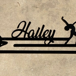 May include: Black metal wall mounted medal holder with the name "Hailey" in a cursive font. Two black silhouettes of dancers are on either side of the name.