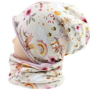 Op de afbeelding: Een crèmekleurige muts en nekwarmer set. De stof heeft een grillige print van vossen, bloemen, regenbogen en eikels. De muts en nekwarmer zijn afgewerkt met een zachte roze stof.
