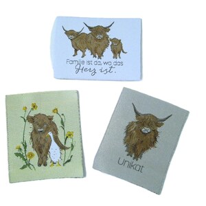 May include: Three fabric patches with Highland cow illustrations. One patch has a family of cows with text, another features a cow with a duck and flowers, and the third has a single cow with the word "Unikat".