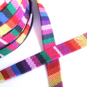 Könnte beinhalten: Eine Rolle aus gewebtem Stoffband mit einem lebendigen, mehrfarbigen Streifenmuster. Das Design besteht aus sich wiederholenden Bändern in Rot, Pink, Lila, Blau, Grün, Gelb und Orange, mit feinen weißen diagonalen Linien über der Textur.